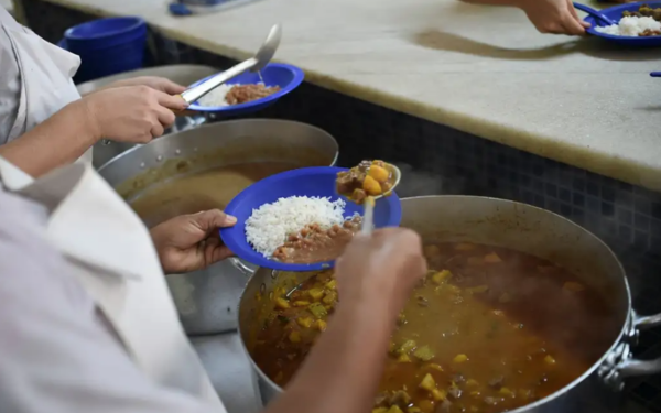 Duas pessoas, de avental branco, estão servindo comida em pratos azuis. Uma delas coloca arroz e feijão no prato, enquanto a outra adiciona um ensopado com legumes. As panelas grandes de alumínio estão cheias de comida quente, e é possível ver o vapor subindo. Ao fundo, outra pessoa segura um prato na fila de distribuição.