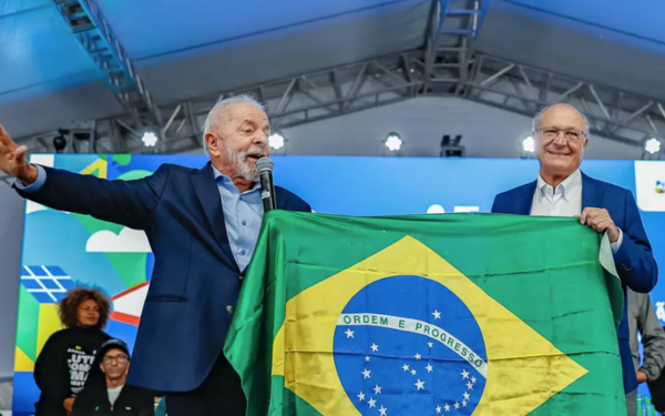 O presidente Luiz Inácio Lula da Silva aparece falando ao microfone com um dos braços estendidos, enquanto segura uma bandeira do Brasil junto ao vice-presidente Geraldo Alckmin, que sorri ao seu lado. Ambos estão sob uma tenda coberta, diante de um palco com iluminação e telão ao fundo. Atrás deles, outras pessoas acompanham o momento com atenção.