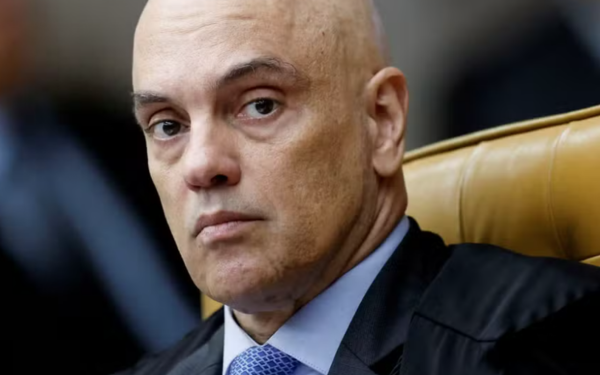 Homem de expressão séria, calvo, de pele clara e vestindo terno escuro com gravata azul clara, está sentado em uma cadeira de couro bege, com encosto alto. A imagem mostra o rosto em destaque, em ambiente fechado, com fundo desfocado.