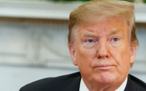 Imagem mostra o ex-presidente dos Estados Unidos, Donald Trump, em um ambiente formal, possivelmente um gabinete. Ele está vestindo terno escuro, camisa branca e exibe expressão séria e concentrada, olhando levemente para o lado. Ao fundo, desfocado, há detalhes decorativos que indicam um espaço institucional.