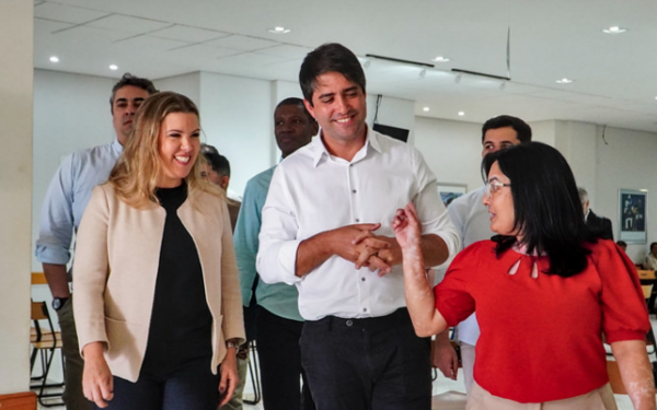 Em um ambiente interno, bem iluminado, com teto branco e luzes embutidas, três pessoas aparecem em destaque no centro da imagem. À esquerda, uma mulher loira, de cabelo solto, veste blazer bege claro e blusa preta, sorrindo enquanto caminha. Ao lado dela, um homem de cabelo escuro e camisa branca sorri enquanto cruza levemente as mãos. À direita, uma mulher de cabelo preto e liso, veste blusa vermelha, calça clara, e fala gesticulando com a mão direita. Ao fundo, outras pessoas observam a interação.