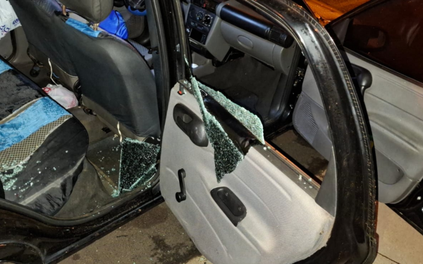 Imagem mostra o interior de um carro com a porta traseira direita aberta e o vidro completamente estilhaçado, indicando arrombamento. Cacos de vidro estão espalhados pelo assento e chão do veículo. O banco traseiro tem capas nas cores preta e azul, e há uma mochila azul visível atrás do banco do passageiro. O ambiente está iluminado artificialmente, sugerindo que a foto foi tirada à noite.