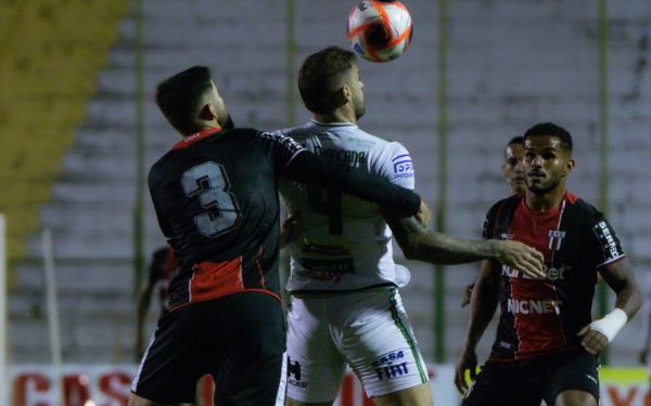 Imagem de uma partida de futebol noturna. Em destaque, dois jogadores disputam a bola pelo alto: um veste uniforme escuro com o número 3 nas costas e o outro, de branco com detalhes verdes, está de costas com a bola próxima à cabeça. A disputa é intensa, com os braços dos atletas se entrelaçando. Ao fundo, outros jogadores observam o lance. A arquibancada aparece vazia e pouco iluminada.