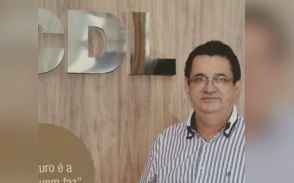 Homem de pele clara, cabelos curtos e escuros, usando óculos e camisa social listrada em tons de branco e azul. Ele está em pé, posando para a foto, com expressão séria e leve sorriso. Ao fundo, uma parede de madeira clara com as letras metálicas “CDL”, sigla da Câmara de Dirigentes Lojistas.