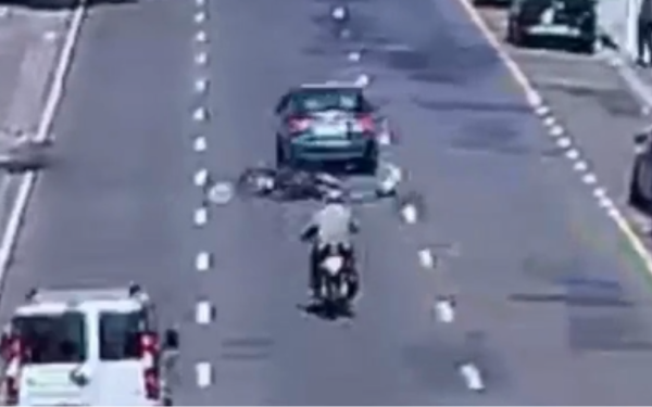 Imagem de uma avenida com três faixas de rolamento. Um carro verde escuro aparece no centro da imagem, momentos após atingir uma motocicleta. A moto e o motociclista estão caídos à frente do veículo. Um outro motociclista, vestindo camisa clara, trafega logo atrás da cena. À esquerda, há uma van branca em movimento. A imagem é de uma câmera de monitoramento urbano.