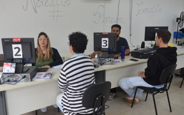 Duas atendentes e um atendente estão sentados atrás de mesas com computadores, atendendo candidatos sentados à frente deles. As estações de atendimento estão numeradas com cartazes "2" e "3". Ao fundo, em uma lousa branca, estão escritas palavras motivacionais como "Acredite", "Persista" e "Conquiste". O ambiente é iluminado e organizado.