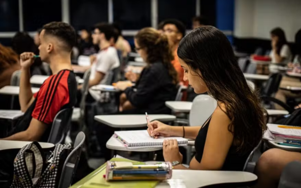 Sala de aula com vários estudantes sentados em carteiras enfileiradas. Em destaque, uma jovem de cabelos longos escreve em um caderno sobre a mesa, concentrada. Ao fundo, outros alunos também acompanham a aula, alguns prestando atenção, outros anotando. A iluminação é artificial e o ambiente lembra uma universidade ou cursinho preparatório.