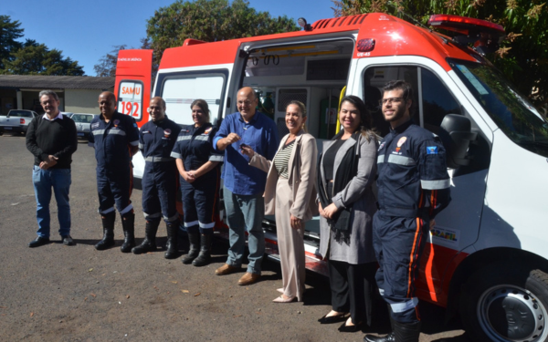 Em um pátio externo, uma equipe do SAMU e representantes da Prefeitura de Franca posam em frente a uma ambulância nova, de cor branca com detalhes em vermelho. O grupo é formado por profissionais uniformizados e autoridades em trajes civis. Um dos representantes segura simbolicamente a chave do veículo, celebrando a entrega da nova viatura.