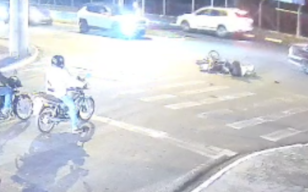 Imagem capturada por câmera de segurança durante a noite. No centro da cena, uma motocicleta e uma pessoa estão caídas no asfalto, indicando um acidente recente. Dois motociclistas observam a situação próximos à faixa de pedestres. Faróis de carros ao fundo iluminam fortemente o cruzamento.