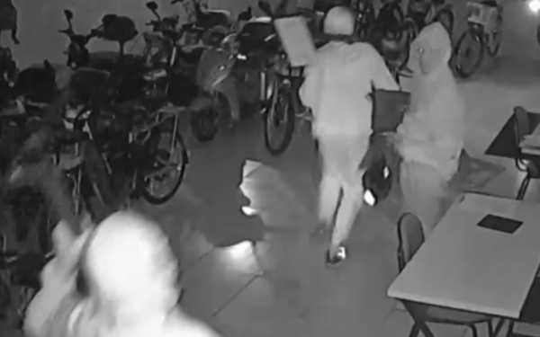 Imagem em preto e branco, capturada por câmera de segurança, mostra o interior de uma loja de patinetes e bicicletas durante a noite. Quatro pessoas encapuzadas, vestindo moletons, aparecem em movimento no local. Uma delas carrega um notebook e outra segura o que parece ser um celular e uma mochila. Ao fundo, há várias bicicletas e patinetes enfileirados. A iluminação é baixa, típica de registros noturnos com visão infravermelha.