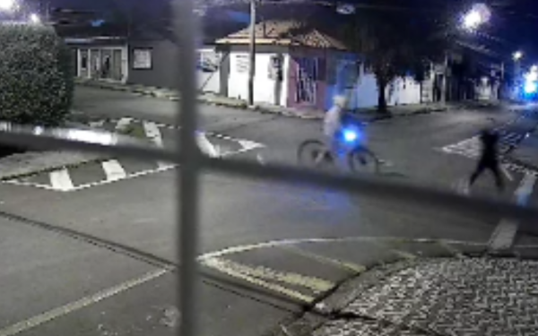 Imagem noturna capturada por câmera de segurança mostra uma rua com pouca iluminação em um bairro residencial. Ao centro, um homem anda de bicicleta com uma lanterna acesa presa na frente, emitindo uma luz azul. À direita, outro homem corre em sua direção. A imagem está parcialmente encoberta por grades de uma janela, indicando que a câmera está dentro de uma residência.