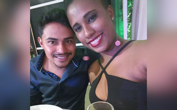 Foto de um casal sorrindo em um ambiente fechado, possivelmente em uma festa. À esquerda, um homem de camisa azul escura com detalhes claros; à direita, uma mulher usando um vestido preto com alças cruzadas e brincos redondos rosas. Ambos olham para a câmera e parecem felizes.