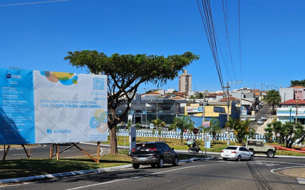 Placa grande de obra da Sabesp instalada em um canteiro gramado na rotatória das avenidas Hélio Palermo e Orlando Dompieri, em Franca. A placa tem fundo azul e branco com os dizeres: “Essa obra vai contribuir para que os rios fiquem mais limpos. Desculpem-nos pelo transtorno”. Ao fundo, aparecem prédios residenciais e comerciais, carros e motocicletas passando pela via, além de tubos laranja e azul utilizados na obra