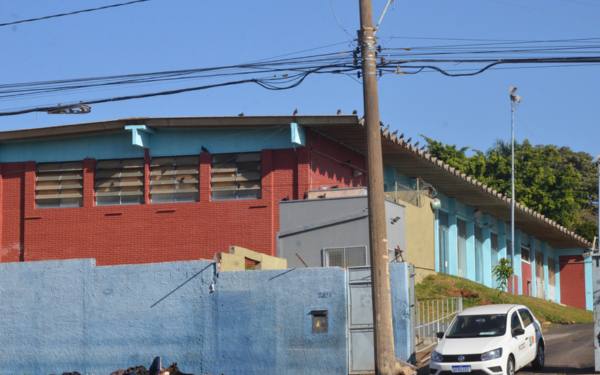 Imagem mostra a fachada de um prédio público com paredes em tons de vermelho, azul e bege. À esquerda, há um muro azul e, ao fundo, janelas com basculantes abertos. Um carro branco oficial está estacionado em frente à entrada lateral do prédio, que fica em uma rua inclinada. Fios elétricos cruzam a parte superior da imagem. É dia e o céu está limpo
