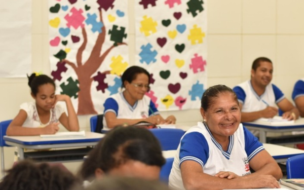 Sala de aula com alunos adultos e idosos sorrindo e atentos durante uma atividade escolar. Ao fundo, há painéis coloridos com desenhos de corações, peças de quebra-cabeça e uma árvore com galhos marrons. As carteiras são azuis e todos os estudantes vestem uniformes brancos com detalhes azuis.