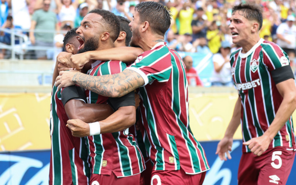 Cinco jogadores do Fluminense comemoram intensamente um gol durante uma partida. Eles vestem o uniforme tradicional do clube, com listras verticais em verde, grená e branco. Um dos atletas está visivelmente emocionado, gritando com força enquanto é abraçado pelos companheiros. Ao fundo, a torcida aparece desfocada, vibrando nas arquibancadas.