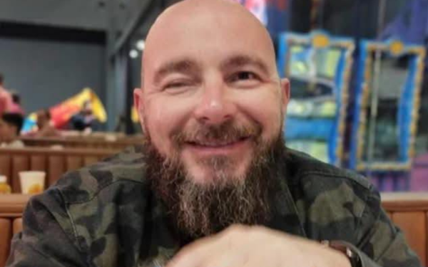 Homem calvo, de barba cheia e escura, sorri enquanto está sentado em um restaurante. Ele veste uma jaqueta camuflada e está com os braços apoiados à mesa. Ao fundo, há luzes coloridas e outras pessoas desfocadas.