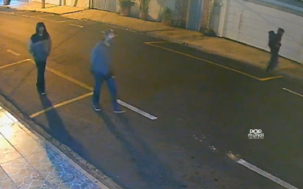 Imagem capturada por câmera de segurança durante a madrugada. Três homens caminham pela rua com tranquilidade. Dois estão no lado esquerdo da imagem, vestindo calça e agasalho com capuz; o terceiro, mais ao fundo, caminha próximo a um portão branco do lado direito. A rua está vazia e iluminada por luz amarelada.