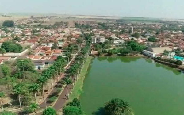 Imagem aérea mostra o lago municipal de Ipuã cheio, com água verde ocupando toda a área. Ao lado, uma avenida margeada por uma fileira de palmeiras e casas de telhado vermelho. Ao fundo, vê-se grande parte da cidade e plantações no horizonte.