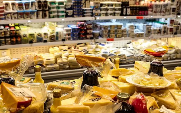 A imagem mostra uma seção de supermercado com prateleiras repletas de queijos e laticínios. Em primeiro plano, há diversos tipos de queijos embalados e dispostos sobre o balcão refrigerado. Ao fundo, as geladeiras exibem mais produtos derivados de leite, como iogurtes e outros queijos.