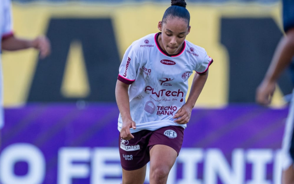 Atleta de futebol feminino, vestindo uniforme branco com detalhes em vinho da Ferroviária, está em campo durante uma partida. Ela aparece concentrada, correndo com a bola, com os olhos fixos para frente e cabelo preso em coque.