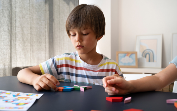 enino com cabelo liso e castanho, usando camiseta listrada com cores claras, está sentado à mesa concentrado enquanto brinca com blocos coloridos de formas geométricas. Ao seu lado, outra criança também participa da atividade, embora apenas o braço dela esteja visível. A mesa é escura e há papéis com desenhos coloridos. Ao fundo, quadros infantis decoram o ambiente iluminado por luz natural.