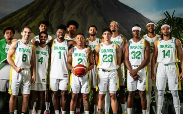 Seleção Brasileira masculina de basquete posa para foto oficial. Os jogadores vestem uniformes brancos com detalhes em verde e amarelo, todos com a palavra Brasil estampada no peito e diferentes números. Ao centro, um atleta segura a bola de basquete. Ao fundo, aparece uma imagem de uma montanha e vegetação, compondo o cenário. O grupo está lado a lado, sorrindo e mostrando união.