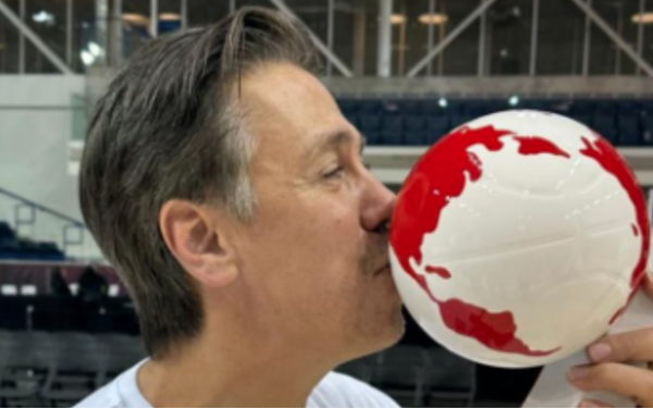 A imagem mostra um homem beijando um troféu em formato de bola de basquete branca com detalhes em vermelho, representando o mapa-múndi. Ele está em um ginásio esportivo, aparentando comemorar uma conquista.