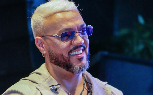 Homem de pele clara, com cabelo loiro platinado e barba bem desenhada, aparece sorrindo. Ele usa óculos de lentes azuis, brinco na orelha esquerda, colar prateado e uma roupa clara com zíper aberto no peito. Ao fundo, o ambiente é escuro e desfocado, sugerindo um local fechado com iluminação azulada.