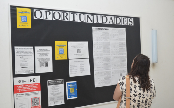 Uma mulher de cabelos escuros e cacheados, vestindo blusa clara estampada, observa um grande quadro de avisos fixado em uma parede branca. No quadro, está escrito em letras grandes a palavra “OPORTUNIDADES” e abaixo dela há várias folhas afixadas com anúncios e informações impressas, algumas com QR Codes para acesso digital.