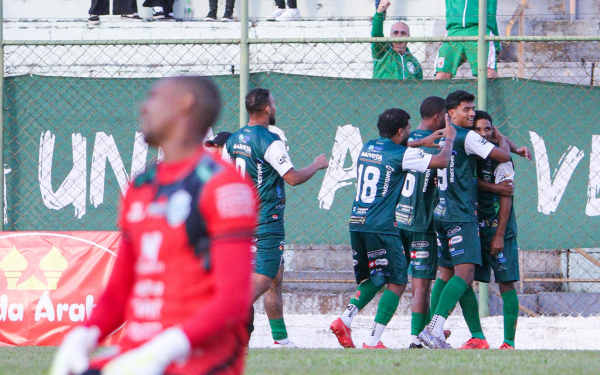 Jogadores da Francana, vestindo uniforme verde, se abraçam e comemoram um gol próximos à lateral do campo, diante de uma grade com faixa verde ao fundo. Um jogador adversário, de uniforme vermelho, aparece desfocado em primeiro plano, transmitindo a frustração.