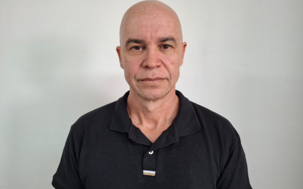 Homem de pele clara, com a cabeça raspada, expressão séria e vestindo uma camisa polo preta. Ele está posicionado de frente, em pé, diante de uma parede branca.