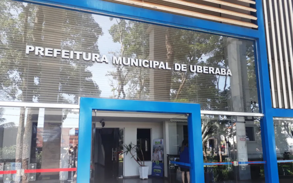 Fachada da Prefeitura Municipal de Uberaba. A imagem mostra a entrada do prédio com estrutura em vidro e moldura azul em destaque. É possível ver o reflexo das árvores na parte superior e algumas pessoas circulando no interior do local.