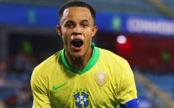 Jogador comemora com expressão de alegria. Ele veste o uniforme amarelo, com o escudo da CBF no peito, detalhes em verde e a braçadeira de capitão no braço esquerdo. Ao fundo, parte do estádio aparece desfocada com torcedores e iluminação.