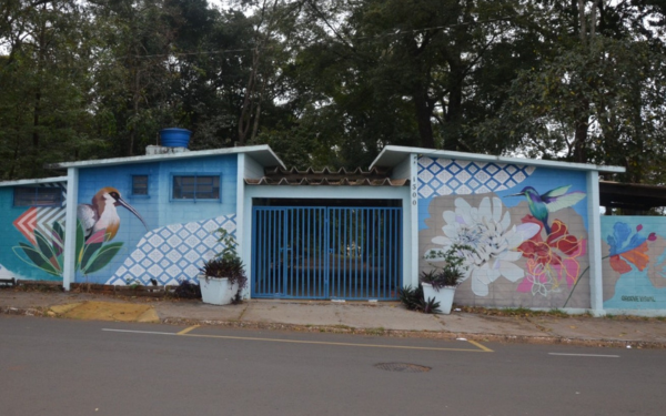 Foto da fachada do Bosque dos Angicos, que é um prédio baixo com paredes pintadas com murais coloridos de pássaros e flores. À esquerda, há um desenho de um pássaro com bico longo, e à direita, um beija-flor em frente a flores grandes e coloridas. O portão central é azul e está fechado. Ao redor, há árvores grandes e uma rua asfaltada em frente ao prédio.