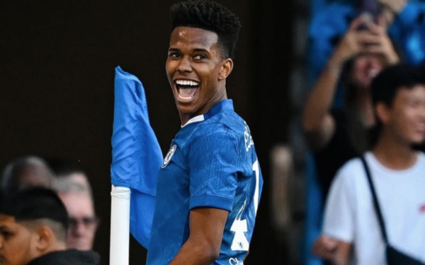 jovem jogador de futebol veste uniforme azul, sorri amplamente e comemora próximo à bandeirinha de escanteio, enquanto torcedores aparecem ao fundo registrando o momento.