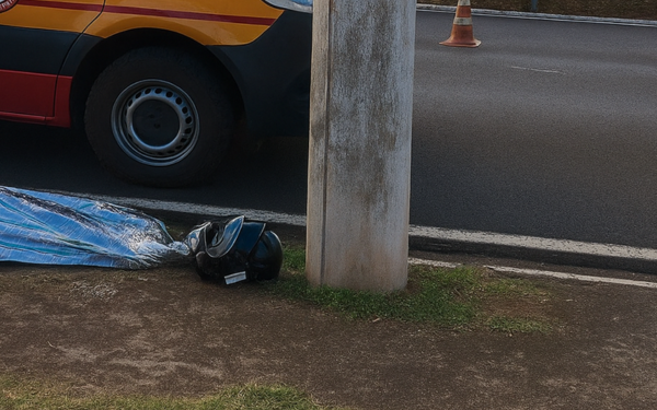 Imagem mostra parte de uma avenida com um poste de concreto no centro do enquadramento. À esquerda, há uma viatura do Corpo de Bombeiros, vermelha com faixas amarelas, estacionada na via. No chão, à frente do poste, está um corpo coberto por um lençol térmico azul, com um capacete preto posicionado próximo à cabeça.