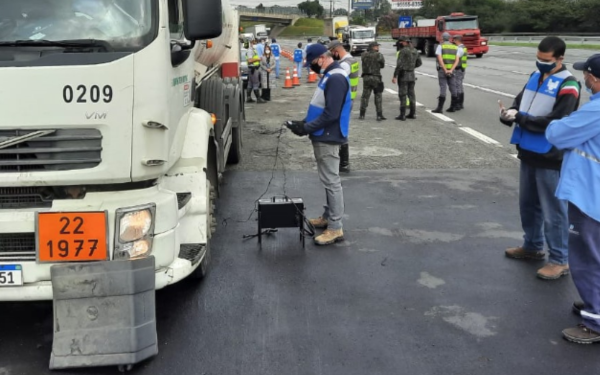 Agentes da Cetesb e da Polícia Militar Rodoviária realizam fiscalização em uma rodovia. Um caminhão está parado no acostamento, enquanto técnicos utilizam equipamentos para medir a emissão de poluentes. Outros veículos e fiscais aparecem ao fundo, acompanhando a operação.
