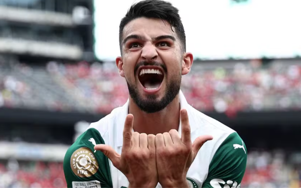 Jogador de futebol com uniforme verde e branco do Palmeiras comemora intensamente um gol. Ele está de boca aberta, gritando com expressão de euforia, e faz um gesto com as mãos, apontando os dedos indicadores e mínimos para cima. No braço, aparece um patch dourado da FIFA no uniforme. Ao fundo, as arquibancadas estão cheias de torcedores.