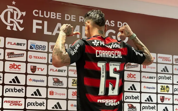 Jogador de futebol veste o uniforme rubro-negro do Flamengo e posa de costas, apontando com os polegares para o nome “Carrascal” estampado na camisa número 15. Ele está diante de um painel com marcas patrocinadoras e o escudo do clube ao fundo.