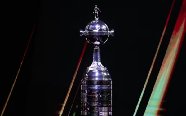 A foto mostra o troféu da Copa Libertadores da América. O objeto é prateado e reluzente, com uma base cilíndrica e uma grande esfera no topo, onde está fixada a escultura de um jogador de futebol em movimento. O fundo é escuro, o que destaca ainda mais o brilho metálico da taça.