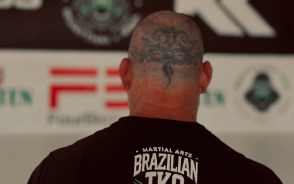 A foto mostra um homem de costas, com a cabeça raspada e uma grande tatuagem na parte de trás do couro cabeludo. Ele veste uma camiseta preta com os dizeres “Martial Arts Brazilian TKO” estampados em branco. Ao fundo, aparecem painéis e logotipos de academias e patrocinadores ligados às artes marciais.