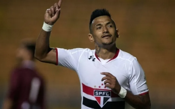 Jogador de futebol com uniforme branco do São Paulo, que tem detalhes em vermelho e preto, aparece comemorando em campo, com o dedo indicador levantado e expressão de confiança.