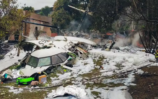 Fotografia mostra os destroços de um avião parcialmente destruído no chão de uma área residencial. O avião está quebrado em várias partes, com a parte frontal visível, incluindo a cabine, onde se lê parcialmente a palavra "Voepass". Espuma branca usada para apagar fogo cobre parte do solo e dos destroços. Ao fundo, fumaça ainda sobe e é possível ver árvores e casas próximas, algumas danificadas pelo impacto.