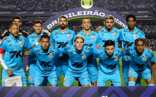 Elenco do Santos Futebol Clube posa para foto oficial antes de partida do Campeonato Brasileiro. Os jogadores vestem uniforme azul claro com detalhes pretos. Eles estão alinhados em duas fileiras: os da frente agachados e os de trás em pé. Ao fundo, vê-se o campo iluminado e o arco com a marca do "Brasileirão".