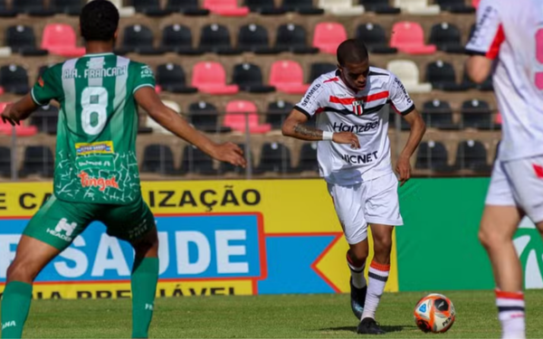 Em campo de futebol, jogador do Botafogo-SP, vestindo uniforme branco com listras horizontais em preto e vermelho, conduz a bola. À sua frente, um atleta da Francana, com uniforme verde e o número 8 estampado nas costas, tenta fechar o espaço. Ao fundo, arquibancadas com cadeiras pretas e vermelhas estão parcialmente ocupadas.