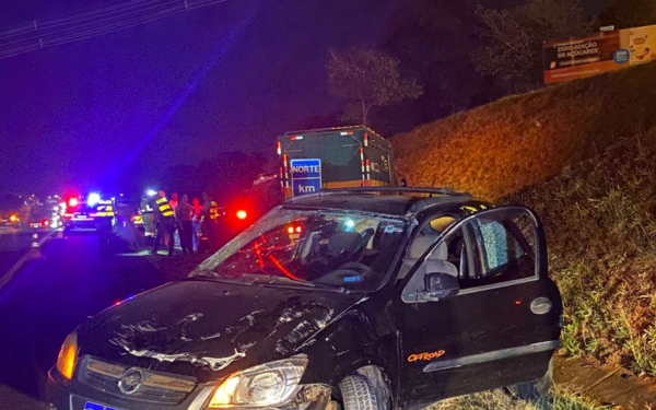 Carro Celta aparece danificado na parte da frente e no teto, indicando que sofreu um acidente. O veículo está parado no acostamento de uma rodovia à noite, com a porta do motorista aberta. Ao fundo, há viaturas da polícia e do resgate com luzes vermelhas e azuis piscando, além de socorristas atendendo a ocorrência.