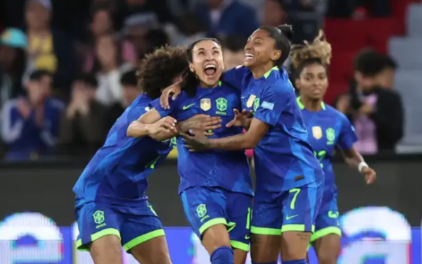 Jogadoras da Seleção Brasileira de Futebol comemoram com empolgação em campo. Elas vestem o uniforme azul com detalhes em verde-limão e exibem expressões de alegria e euforia após um gol ou vitória. Uma das atletas está no centro, sendo abraçada pelas companheiras, enquanto o público aparece desfocado ao fundo.