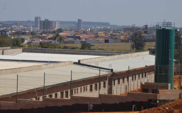 Fotografia mostra a construção do novo prédio do Núcleo de Gestão Assistencial (NGA) em andamento. A estrutura, de grande porte, tem paredes em alvenaria e telhado metálico já instalado. Ao lado, há um reservatório de água cilíndrico, pintado de verde. O cenário de fundo revela parte da cidade, com casas e prédios ao horizonte.