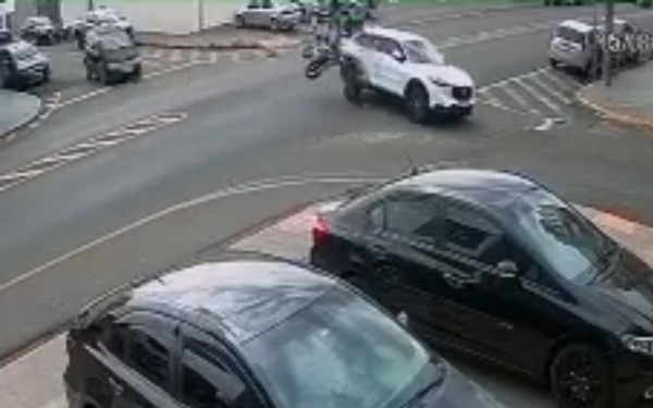 Imagem de uma câmera de segurança mostra o exato momento de um grave acidente de trânsito em um cruzamento urbano. Um carro branco, que avança a sinalização de parada obrigatória, colide com uma motocicleta que seguia pela via preferencial. A força do impacto faz com que o motociclista seja lançado para o alto junto com a moto. No primeiro plano, aparecem dois carros estacionados. A via é asfaltada, com faixas de sinalização visíveis no chão.